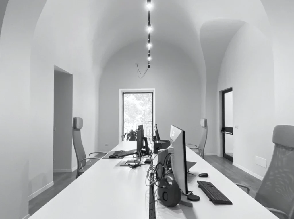 Uno spazio ufficio moderno progettato dal Team Ipogeo Architetti è caratterizzato da un lungo tavolo bianco, diversi computer e sedie con schienale alto. La stanza vanta un soffitto alto con luci sospese e grandi aperture ad arco, che consentono alla luce naturale di entrare attraverso l'ampia finestra all'estremità. Ipogeo Architetti