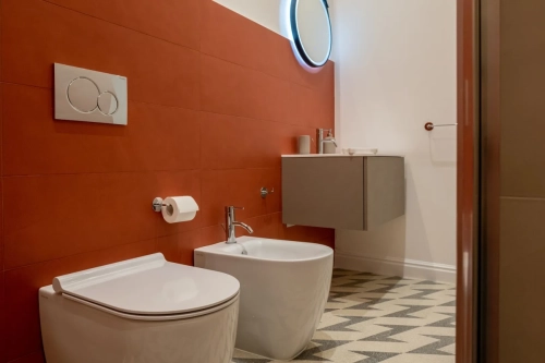 Bagno moderno con piastrelle rosse alle pareti, con un water e un bidet montati, un lavandino a parete con un rubinetto, uno specchio rotondo retroilluminato e un pavimento con motivi geometrici. Rotolo di carta igienica montato a parete e un anello portasciugamani sulla parete adiacente. Ipogeo Architetti