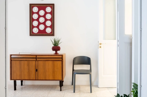 Un ingresso minimalista presenta una credenza in legno sormontata da una pianta in vaso e una ciotola decorativa di frutta rossa. Sopra la credenza è appesa un'opera d'arte incorniciata con motivi circolari su uno sfondo rosso. Sulla destra, una sedia grigia è appoggiata a una porta bianca. Ipogeo Architetti