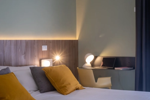 Una camera da letto moderna è dotata di un letto con cuscini gialli e grigi e biancheria da letto bianca. La parete dietro il letto ha un pannello di legno con illuminazione integrata. Una scrivania nell'angolo contiene un computer portatile, un libro e una lampada dal design unico. La stanza ha un'atmosfera accogliente ed elegante. Ipogeo Architetti