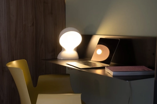 Uno spazio di lavoro moderno è caratterizzato da una lampada da scrivania elegante, un laptop aperto che riflette la luce e un libro chiuso. La scrivania ha una superficie scura contro un mix di legno e pareti color pastello. Una sedia gialla è posizionata sulla scrivania. L'illuminazione crea un'atmosfera calda e invitante. Ipogeo Architetti