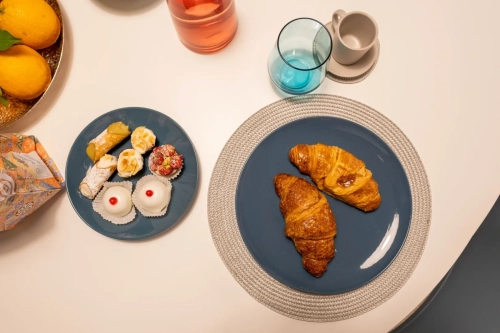 Un tavolo con un piatto rotondo di pasticcini assortiti e un piatto più grande con due croissant. Vicino, c'è una piccola tazza, un bicchiere bluastro su una tovaglietta intrecciata, una bottiglia con un liquido rosa-rossastro e una ciotola con delle arance. Ipogeo Architetti