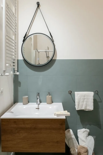 Un bagno moderno con uno specchio circolare appeso sopra un mobile lavabo in legno. La zona lavabo ha dei dispenser di sapone beige, un porta spazzolino e una saponetta. Un asciugamano è appeso a un portasciugamani sulla destra, e delle pantofole bianche e un asciugamano beige sono sul pavimento. Ipogeo Architetti