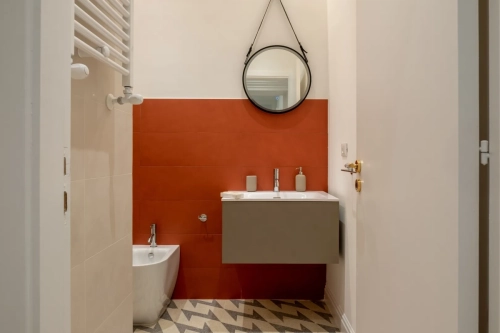 Un bagno moderno con pareti piastrellate di rosso, uno specchio rotondo sopra un lavandino a parete e un bidet sulla sinistra. Il pavimento presenta un motivo di piastrelle a zig zag in grigio e bianco. Pareti bianche e una porta sono parzialmente visibili sulla destra. Ipogeo Architetti