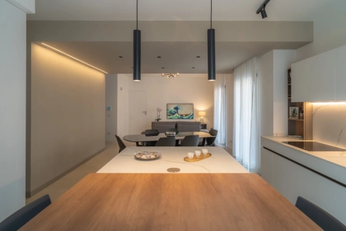 Moderna zona soggiorno e pranzo open space con design minimalista. Presenta un tavolo da pranzo in legno in primo piano, luci a sospensione, un'accogliente zona soggiorno con TV e ampie finestre con tende trasparenti che illuminano lo spazio. Ipogeo Architetti