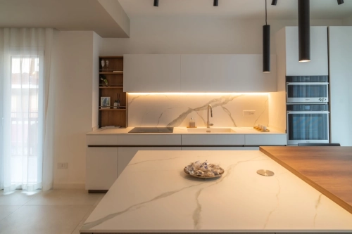 Cucina moderna con ripiani in marmo bianco e illuminazione integrata. Dispone di piano cottura, forno e lavello incorporati. Un piatto con degli spuntini è sul bancone dell'isola. Luci pendenti scure pendono dal soffitto e tende trasparenti coprono una finestra. Ipogeo Architetti