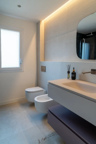 Un bagno moderno con un water e un bidet a parete, un grande specchio sopra un mobile lavabo sospeso con un lavandino a catinella e una finestra che lascia entrare la luce naturale. La stanza è illuminata da luci da incasso e luci ambientali sopra lo specchio. Ipogeo Architetti