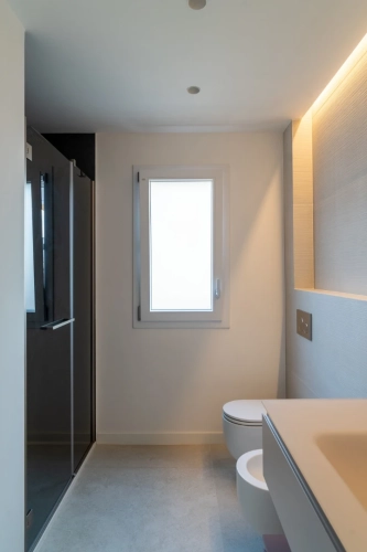 Un bagno minimalista con una finestra smerigliata, una doccia con cabina in vetro sulla sinistra, un water a parete e un lavandino incorporato con illuminazione ambientale sulla destra. Pareti e pavimenti dai toni neutri creano un aspetto pulito e moderno. Ipogeo Architetti