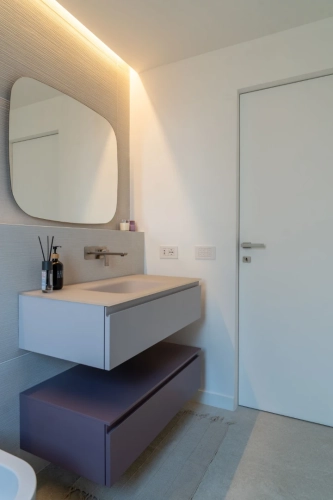 Bagno moderno dal design minimalista. Presenta un grande specchio, un elegante lavandino a parete e sanitari contemporanei. L'illuminazione ambientale e la combinazione di colori neutri creano un'atmosfera serena. Una porta è visibile sulla destra. Ipogeo Architetti