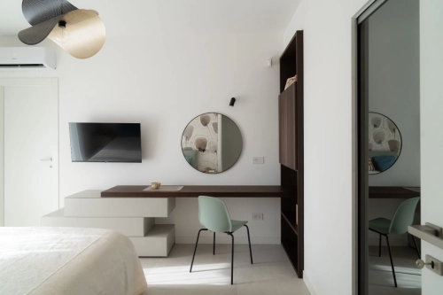 Camera da letto minimalista con TV a parete, specchio rotondo sopra la scrivania e sedia verde chiaro. Toni neutri e uno stile di arredamento moderno creano un'atmosfera serena. Una porta con specchio a figura intera e un'esclusiva luce a soffitto completano lo spazio. Ipogeo Architetti