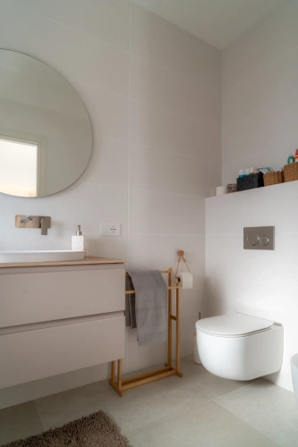 Bagno minimalista con specchio rotondo sopra un lavandino con mobiletto, dotato di rubinetto a parete. Un asciugamano è appeso a un supporto di legno accanto a un water a parete. Gli scaffali contengono articoli da toeletta in cestini intrecciati. Piastrelle di colore chiaro ricoprono le pareti e il pavimento. Ipogeo Architetti