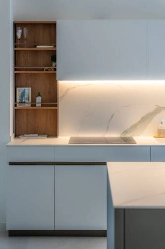 Interno di una cucina moderna con mobili bianchi e ripiani in marmo. Ci sono ripiani in legno aperti con bicchieri e una piccola pianta. Un piano cottura a induzione è integrato nel bancone con una sottile illuminazione sottopensile che illumina il paraschizzi. Ipogeo Architetti