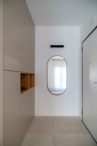 Ingresso moderno e minimalista con mobili beige chiaro con mensole aperte, un grande specchio ovale sulla parete che riflette una finestra con tende trasparenti e una porta bianca. Il pavimento è piastrellato in grigio chiaro. Ipogeo Architetti