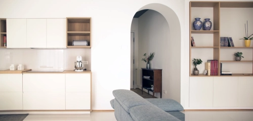 Un soggiorno accogliente e minimalista con pareti bianche e un design pulito. Lo spazio presenta una zona cucina sulla sinistra, un divano grigio in primo piano, una porta ad arco centrale che conduce a un corridoio e mensole integrate sulla destra che espongono libri e decorazioni. Ipogeo Architetti