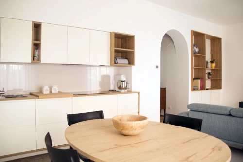 Una cucina e sala da pranzo moderne con mobili bianchi con accenti in legno, un tavolo da pranzo rotondo in legno con una ciotola coordinata, sedie nere, una porta ad arco, scaffali aperti con decorazioni e parte di un divano blu visibile sulla destra. Ipogeo Architetti