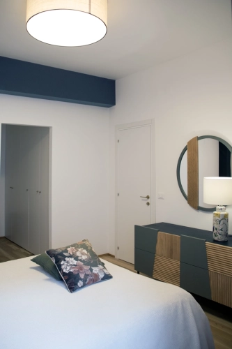 Una camera da letto moderna presenta un letto bianco con cuscini floreali, una cassettiera blu con pannelli in legno e uno specchio rotondo sopra, e una lampada da tavolo bianca con una base a motivi. La stanza ha pareti bianche, una striscia decorativa blu vicino al soffitto e una porta che conduce a un altro spazio. Ipogeo Architetti