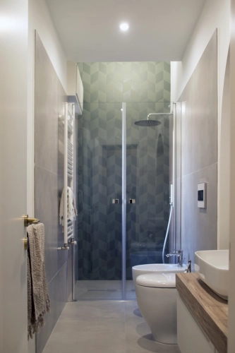 Un bagno moderno con una doccia in vetro con piastrelle verdi geometriche. Lo spazio comprende un water a parete, un lavandino bianco su un mobiletto in legno chiaro e un asciugamano appeso a una barra accanto a una porta con una maniglia dorata. Il design generale è elegante e contemporaneo. Ipogeo Architetti