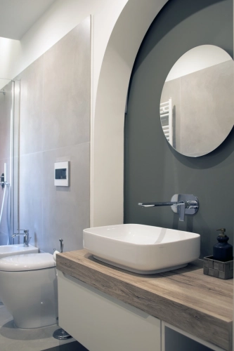 Un bagno moderno presenta un design minimalista. Include un lavandino bianco rettangolare con un piano di lavoro in legno, uno specchio circolare sopra il lavandino, un water e un bidet bianchi e sanitari contemporanei. Le pareti sono un mix di piastrelle grigio chiaro e vernice verde scuro. Ipogeo Architetti