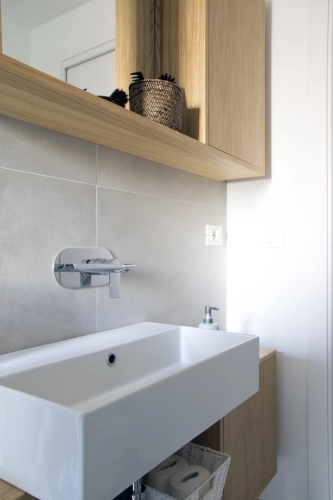 Bagno moderno con lavandino rettangolare bianco, rubinetto a parete e dispenser per sapone. Mobiletto in legno chiaro sopra con cestino portaoggetti e pareti piastrellate grigio chiaro. Design minimalista con linee pulite e accenti in legno naturale. Ipogeo Architetti