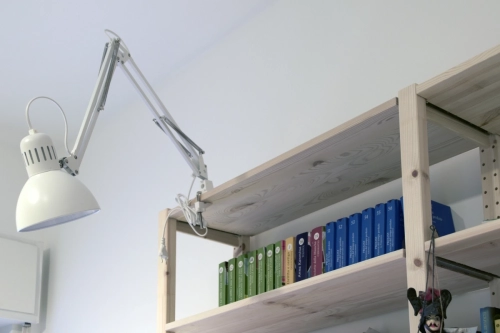 Una lampada da scrivania bianca regolabile è fissata a uno scaffale di legno, inclinata verso il basso. Lo scaffale sottostante contiene una fila ordinata di libri colorati, con volumi verdi, blu e alcuni bianchi visibili. Lo sfondo è un semplice muro bianco. Ipogeo Architetti