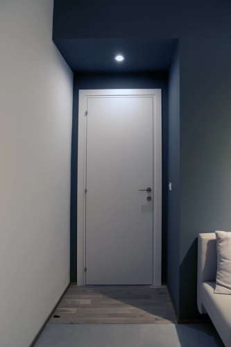 Un corridoio minimalista con pareti e soffitto scuri, con una porta bianca chiusa. Un faretto illumina l'area sopra la porta, mentre a destra è visibile il bordo di un divano di colore chiaro. Il pavimento è costituito da piastrelle grigio chiaro e scuro. Ipogeo Architetti