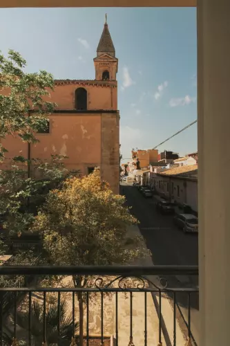 Una vista da un balcone cattura una strada illuminata dal sole con auto parcheggiate, fiancheggiata da edifici storici. In evidenza, una chiesa con un alto campanile appuntito si erge sulla sinistra. Alberi verdi aggiungono un tocco di natura alla scena urbana sotto un cielo azzurro e terso.