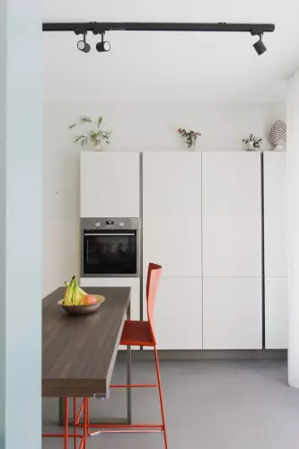 Cucina minimalista con mobili bianchi e forno incorporato. Un'isola in legno con una fruttiera e seggioloni arancioni fornisce posti a sedere. Il soffitto è caratterizzato da un moderno binario di illuminazione nero e piccole piante e oggetti decorativi adornano la parte superiore dei mobili.